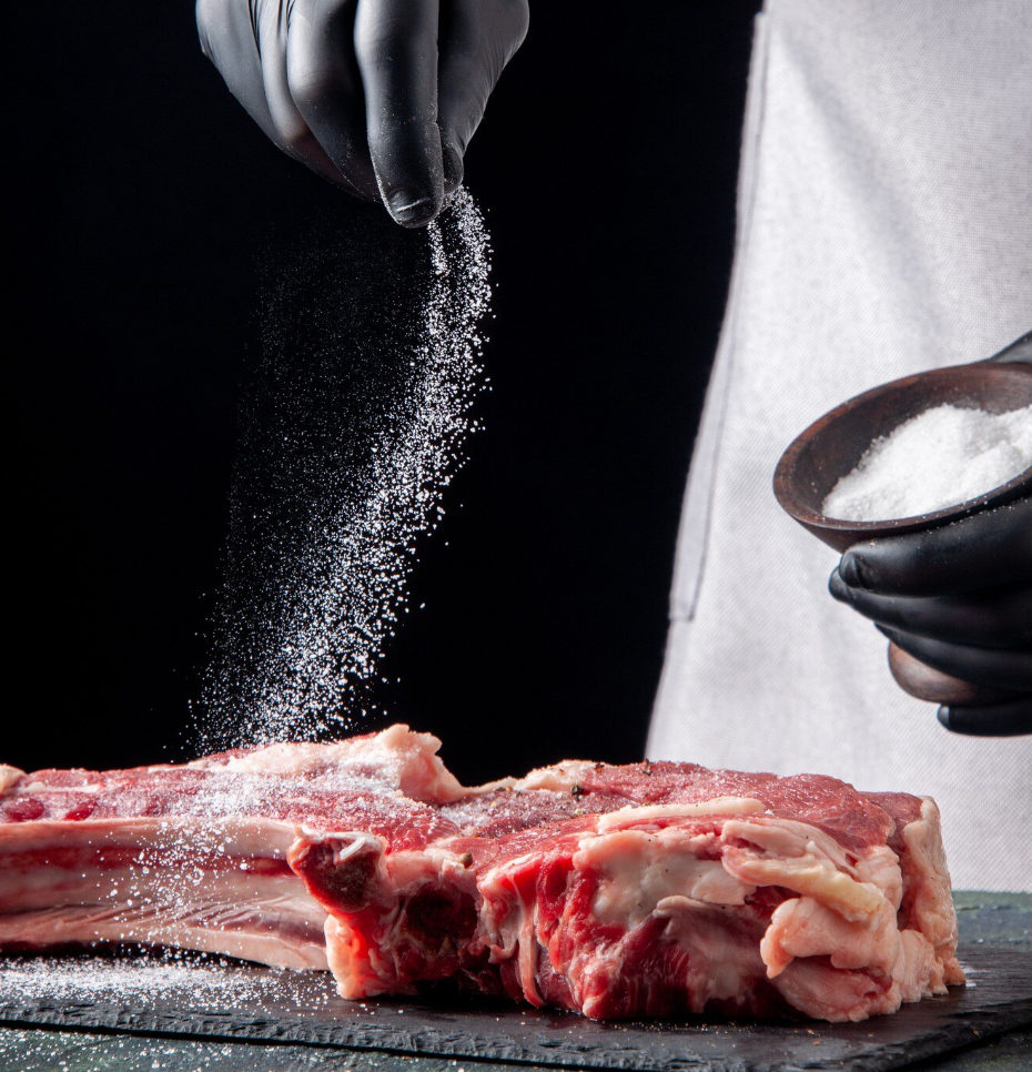 A chef seasoning a large steak with coarse salt